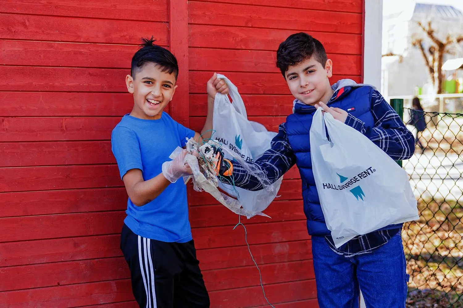 Två glada unga pojkar står framför en röd fasad och visar upp att de har plockat skräp och deras plastpåsar från organisationen Håll Sverige Rent