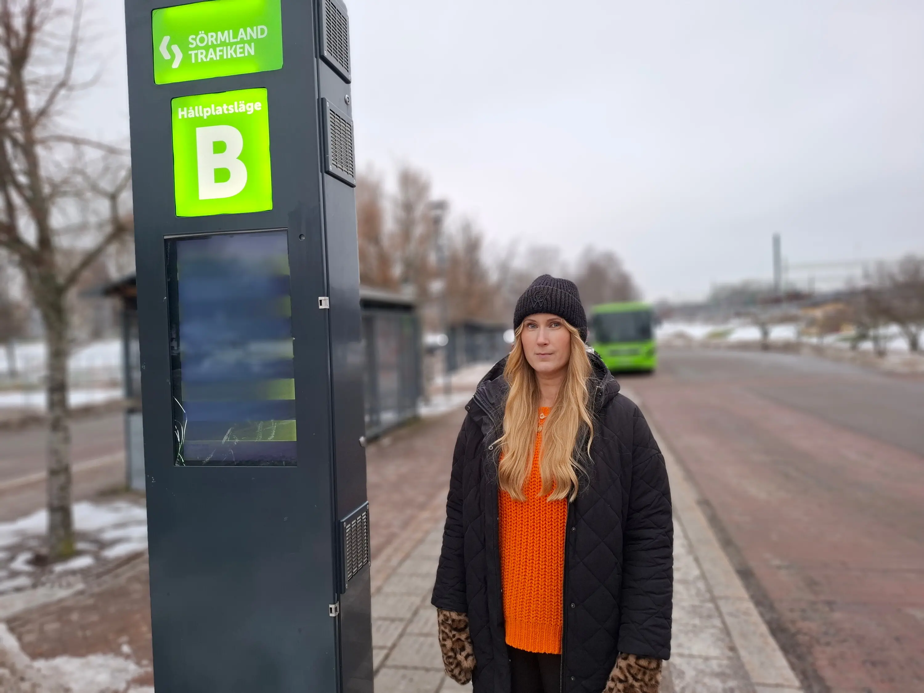 En person står bredvid en skylt med texten "Sörmland Trafiken" och "Hållplatsläge B". En buss syns i bakgrunden.