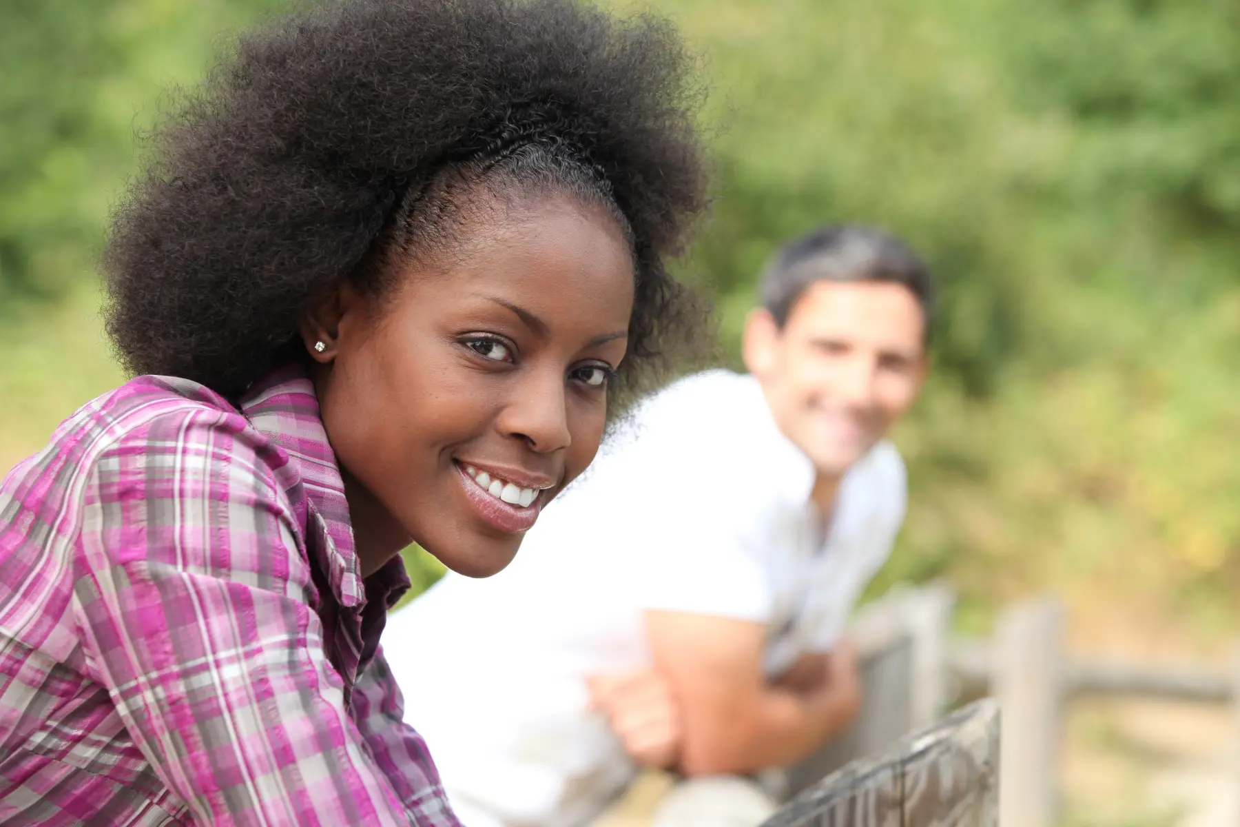 En mörkhyad kvinna ler in i kameran, en man står i bakgrunden och syns oskarpt. 