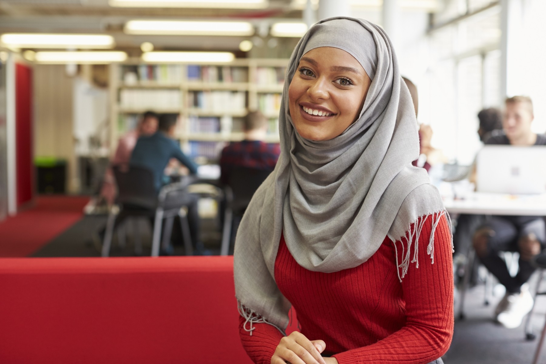 Leende ung kvinna i grå slöja och röd tröja sitter i ett bibliotek eller en studiemiljö.