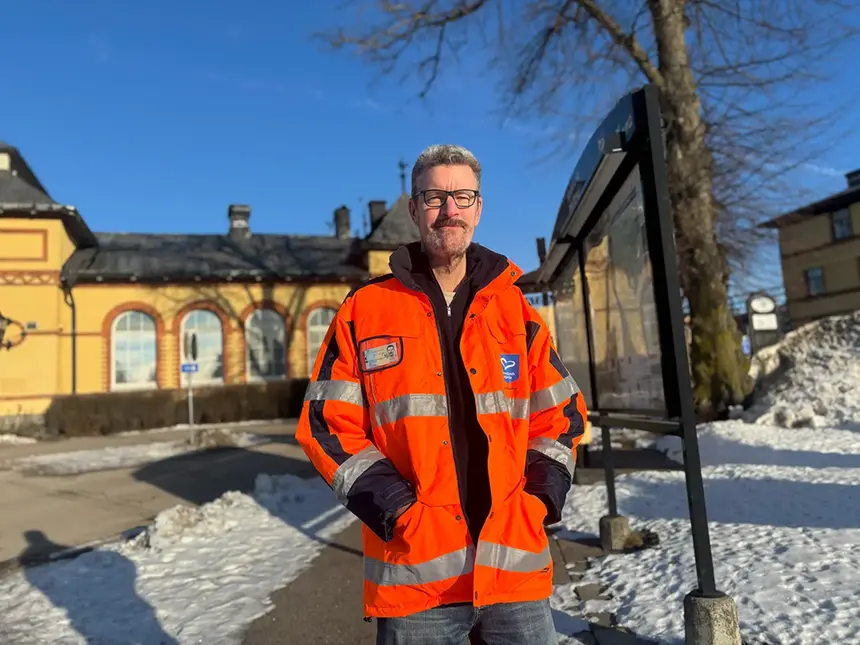 En man i varselväst med texten "’SÄKER’" står framför en byggnad och en busshållplats. Marken är snöig och det är blå himmel.